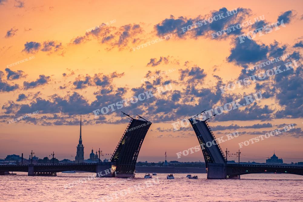 На фотографии изображен разведенный Дворцовый мост на фоне заката в Санкт-Петербурге. На заднем плане виднеется Петропавловская крепость. Фотография идеально подходит для использования в туристических материалах, статьях о Санкт-Петербурге и в промо-материалах о белых ночах.